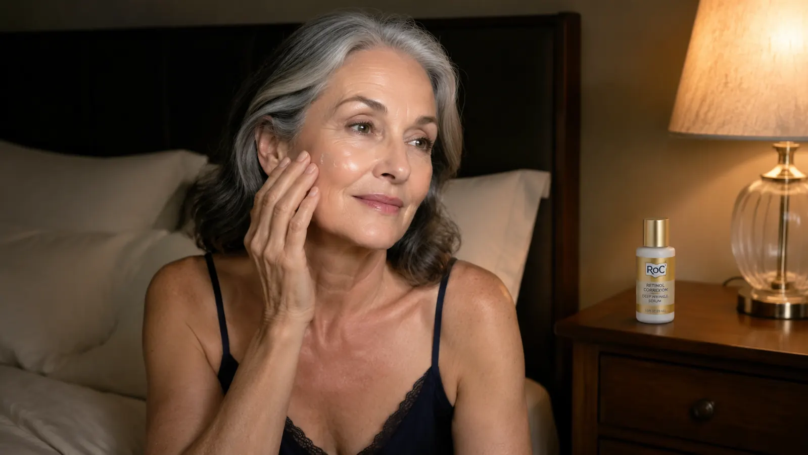 Mature woman applying a small amount of serum to her cheek before bed in dim bedroom lighting